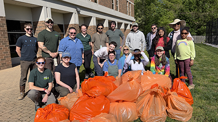 Large group photo outside a building. 18 individuals smiling facing the camera with orange trash bags filled up after a days work of cleaning. 