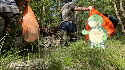 Two men walking away from the camera holding orange trash bags. Stewart the Turtle is in front of one of the trash bags smiling while facing the camera. 