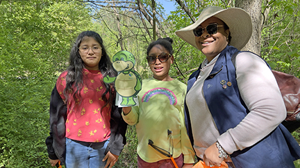 Three women - the one in the center holding a cutout of Stewart the Turtle - smiling at the camera after helping pick up trash.