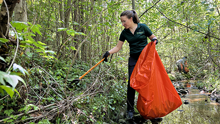 Woman holding an orange trash bag and a reach extender cleaning up trash along a creek bed. 