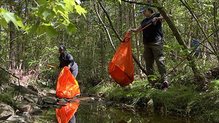 Two men holding orange trash bags cleaning up trash by a creek in the woods