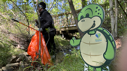 Woman facing away and to the left side of the photo holding an orange bag and reach extender to collect trash. Stewart the Turtle is to the right of the screen smiling looking into the camera. 