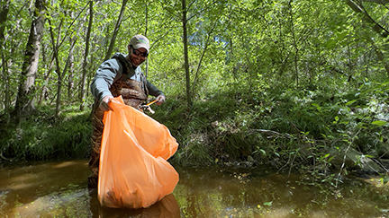 Man holding an orange trash bag picking up trash in a creek in the woods. 