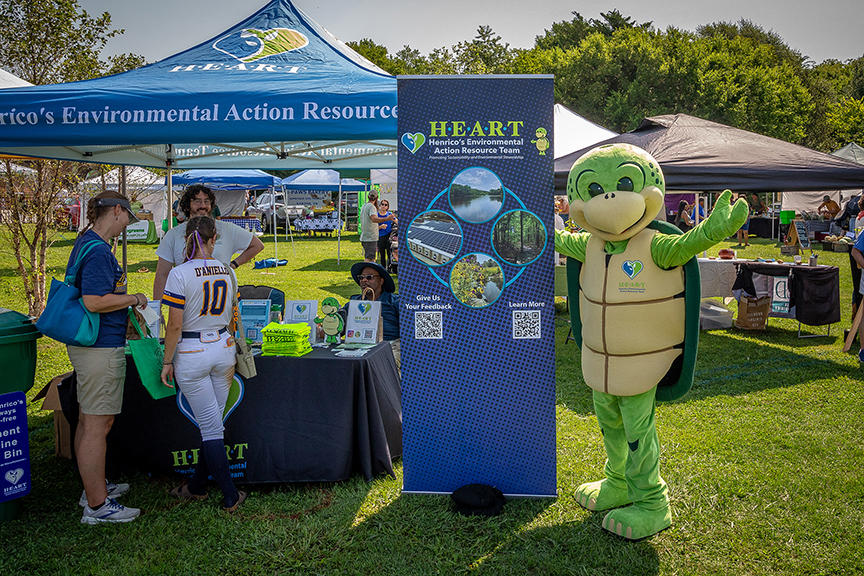 Life size Stewart the Turtle mascot standing next to the H.E.A.R.T banner smiling with his hands up with another H.E.A.R.T tent to his left and people standing in front of the tent. 