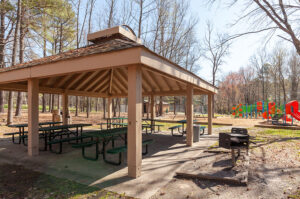 Photograph of a shelter with green picnic tables, small grills, and a playground in the background.