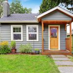 Small grey house with wooden deck. Front yard with flower bed and lawn