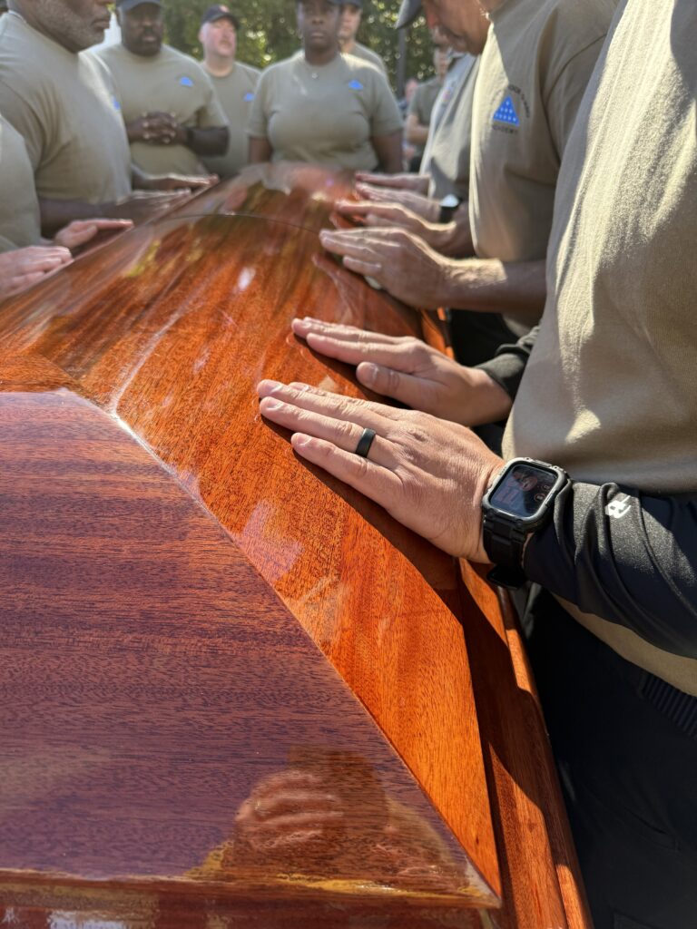 As part of an honor guard training, participants place their hands on a wooden casket.