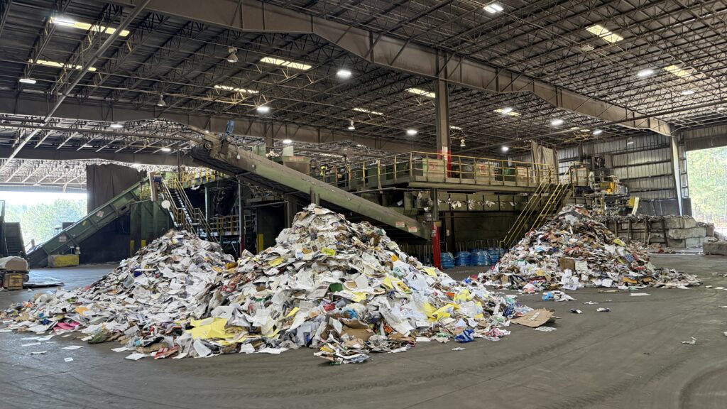A large indoor recycling facility with piles of sorted paper and cardboard. Conveyor belts and industrial equipment are visible, facilitating the recycling process. The roof is supported by metal beams, and lighting fixtures illuminate the area.