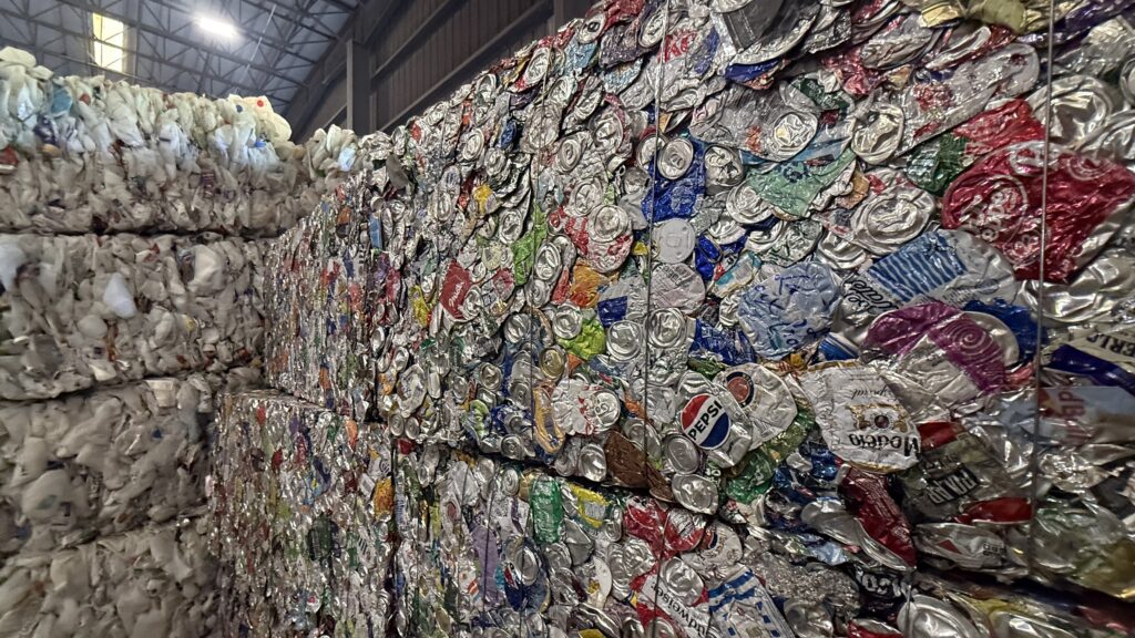 Stacks of compressed recyclables, including a wall of crushed aluminum cans featuring recognizable brands like Pepsi and Coca-Cola, are organized in a large industrial facility. The ceiling is high, and sunlight filters through overhead.