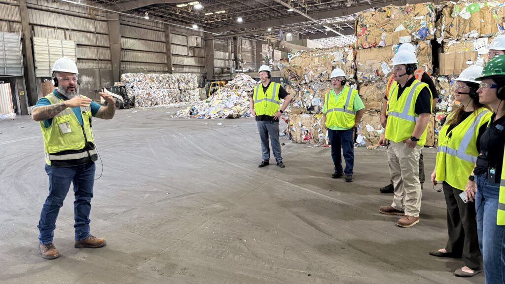 A group of people in safety vests and helmets are gathered in a recycling facility. One person appears to be speaking or gesturing to the others. Large stacks of sorted recyclable materials, including cardboard and plastics, are visible in the background.