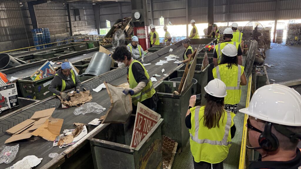 People wearing safety vests and helmets work along a conveyor belt in a recycling facility. They are sorting various materials like cardboard and paper. The facility is spacious, with high ceilings and various equipment around.