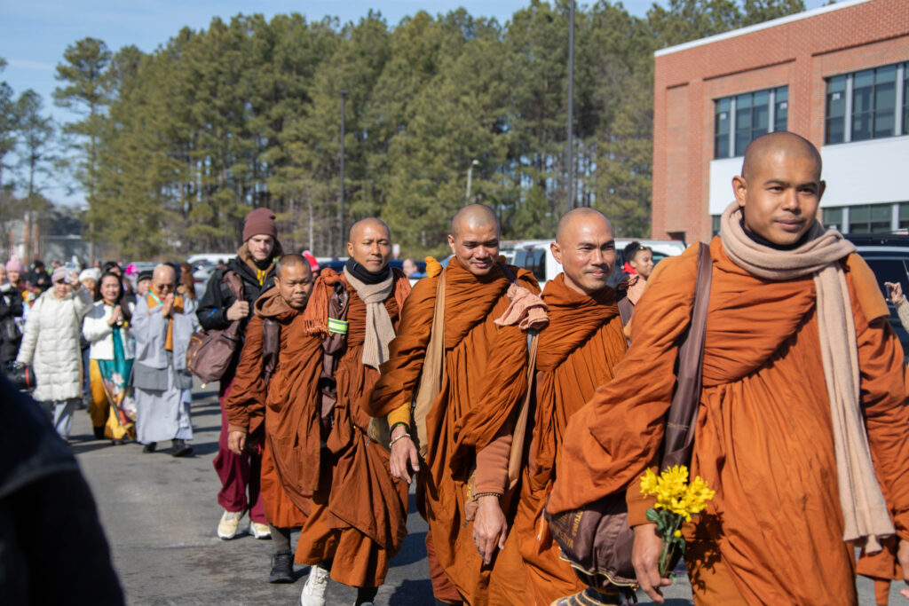 Buddhist monks in orange robes walk single file holding flowers.