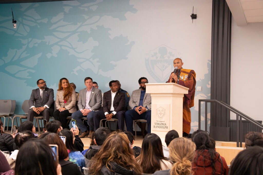 10 adults sit on stage and listen to a monk speak at a podium.