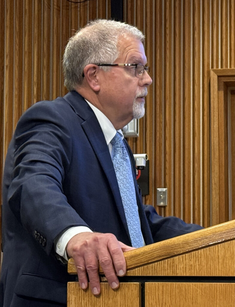 A man in a suit speaks at a lectern in a meeting room.