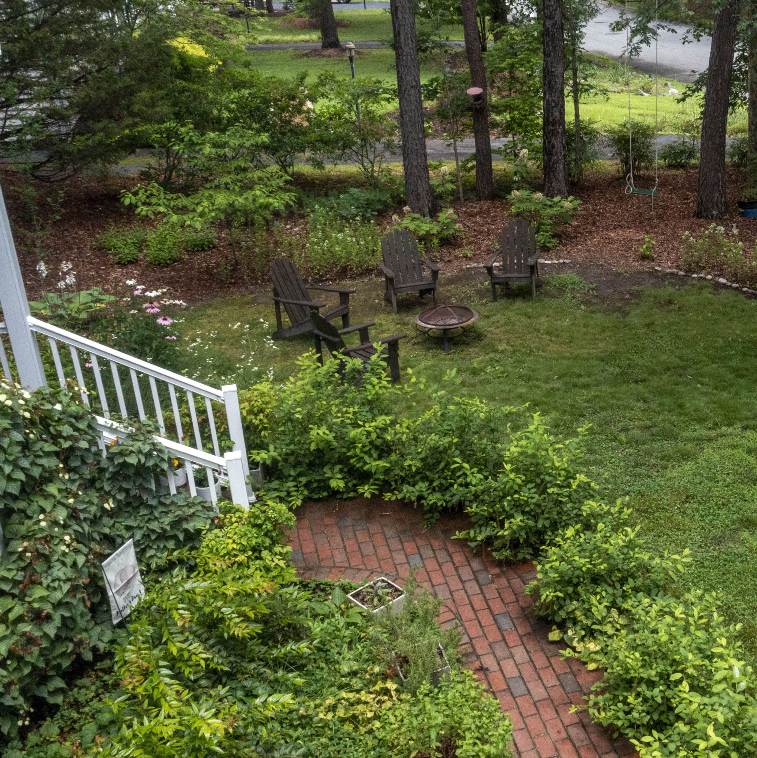 photo of a lush front yard with brown lawn chairs pink flowers and a brick walkway lined with native shrubs.