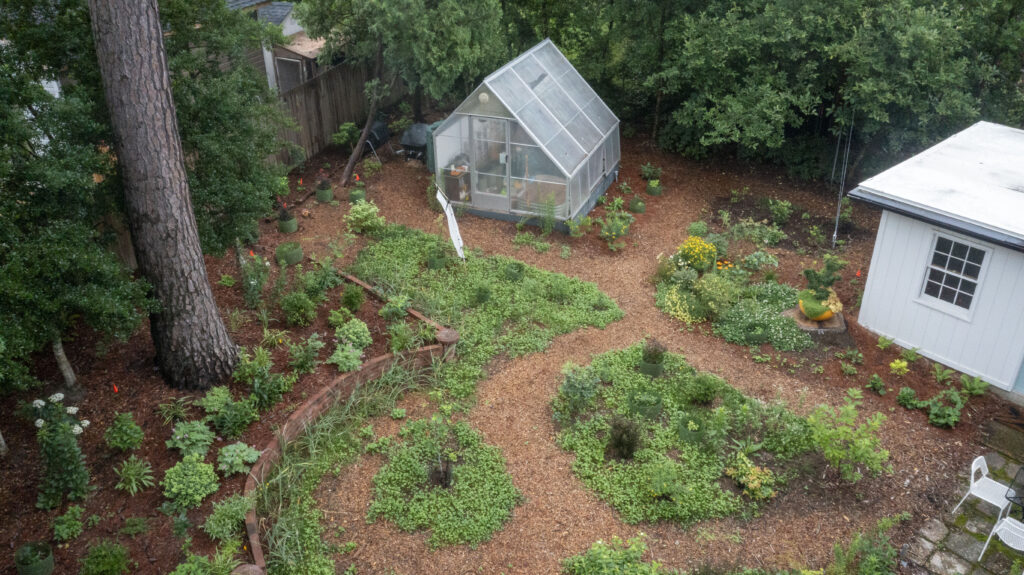 aerial photo of a backyard with much of the space taken up by garden beds and paths made up of pine needles. There is also a greenhouse and compost bin in the yard.