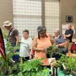 Event attendees browse various plants on a table, inspecting the label descriptions.