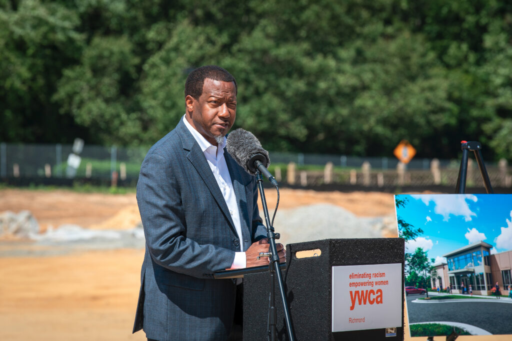 A person is speaking at a podium with a YWCA Richmond sign, set outdoors with trees and a construction site in the background. A display board shows an illustration of a building.