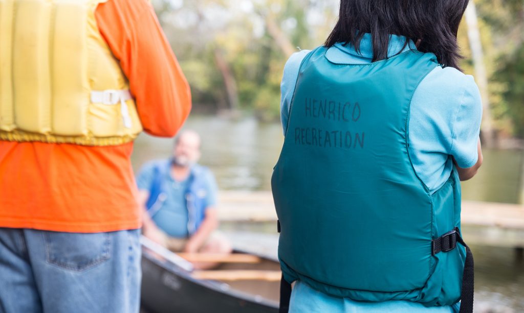 Two people in PFDs listening to instructions from a man sitting in a canoe.