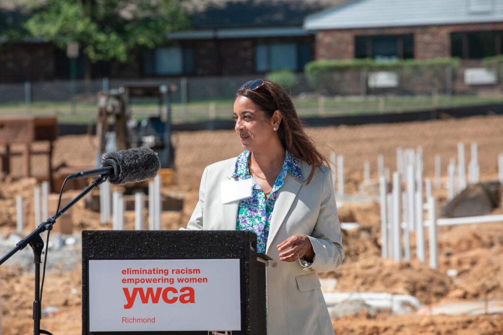 An individual is speaking at a podium with a "ywca" logo, which reads "eliminating racism empowering women" and mentions Richmond. The background shows a construction site with pipes and machinery.