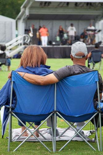 Photo of entertainers on a stage with the focus on two event guests sitting together in folding chairs on the lawn.