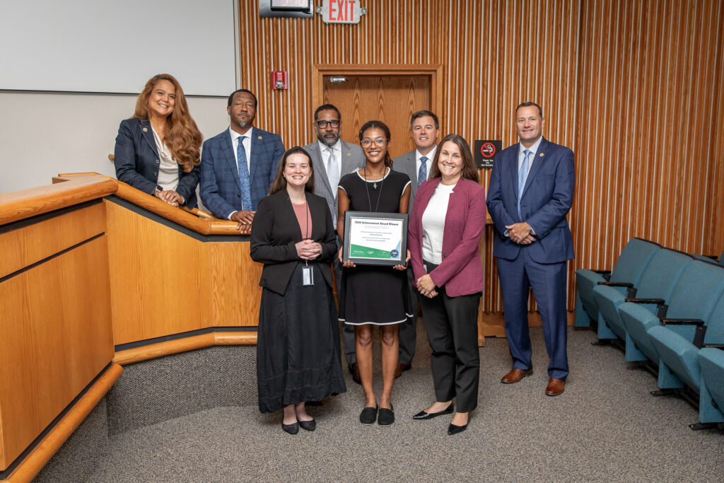 A group of people stand in two rows and the person in the middle holds a framed award.