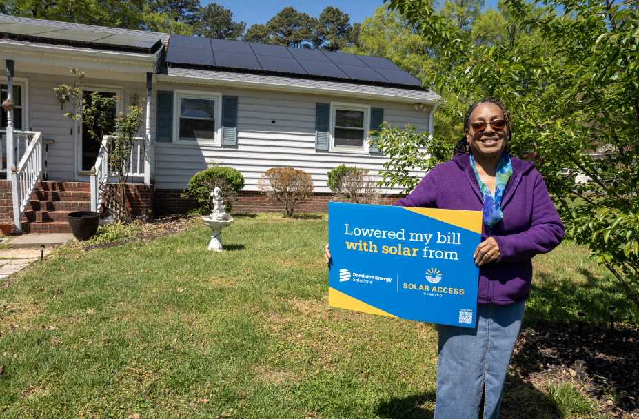 A woman holding a blue sign that advertises Solar Access Henrico and Dominion Energy Solutions stands in front of a house with solar panels on the roof.