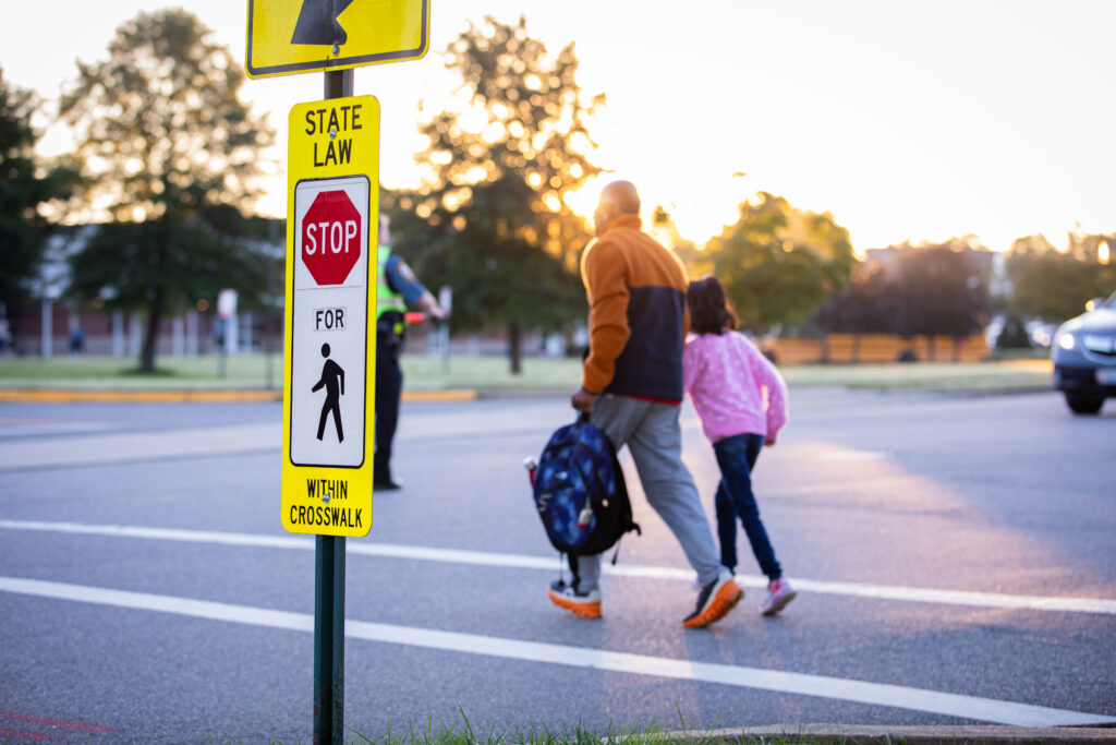 A man and child walk on a crosswalk by a sign advising motorists to stop.
