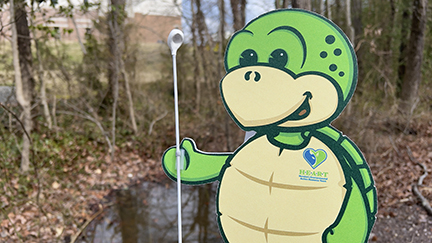 Turtle mascot holding a white mosquito scoop against winter woods & a small bit of water