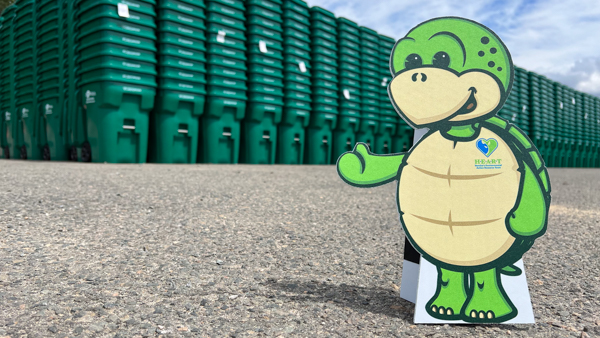 Stewart the turtle mascot cutout in front of tall stacks of green recycle bins with white clouds & blue sky 