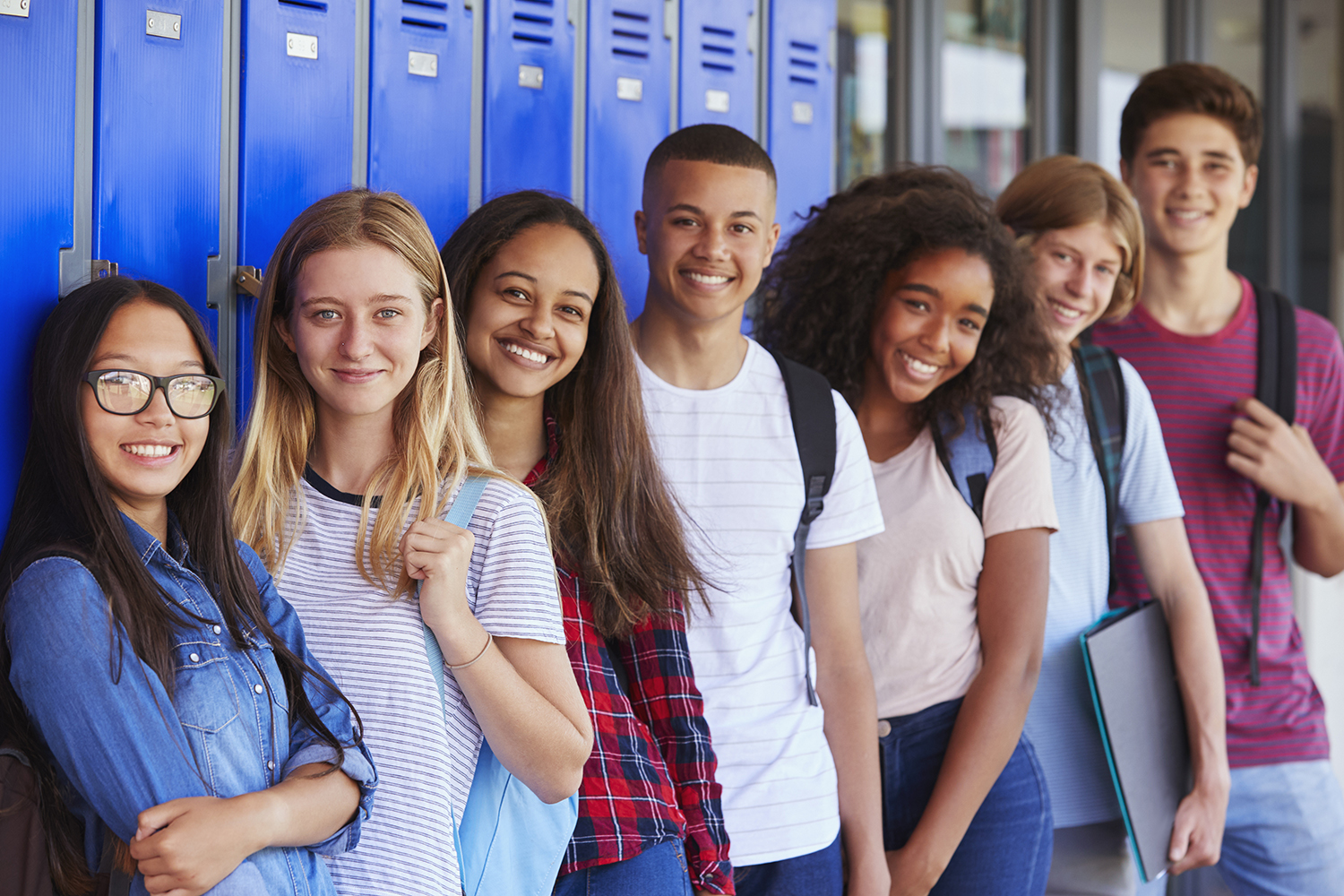 Diverse group of seven smiling teens standing next to blue lockers teens