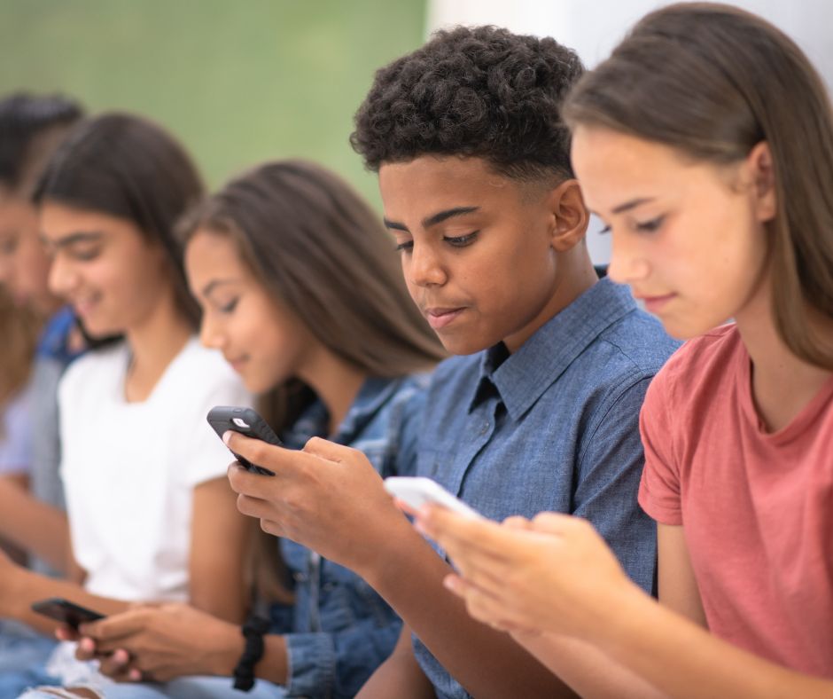 Group of diverse teenagers sitting side by side, each focused on their smartphones in a casual setting.