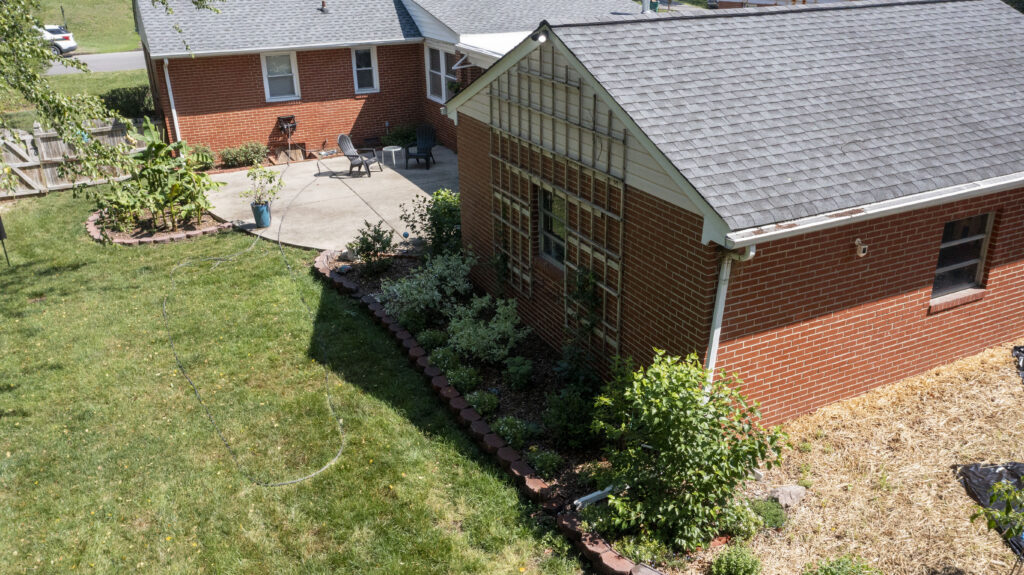 An aerial view of the back of a house with well-landscaped plants in separate plots along the back of a home. A trellis hangs on the back of the house for climbing plants.