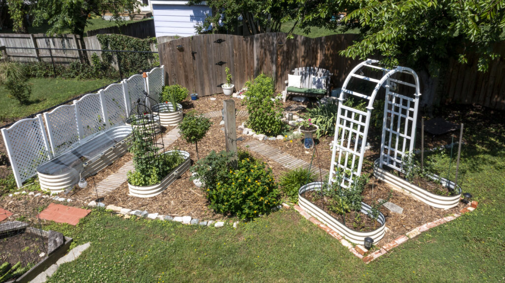 A portion of a yard without turf grass, filled with raised garden beds and an intentional path to visit the various plants. A white arch sits at the entrance to the garden.