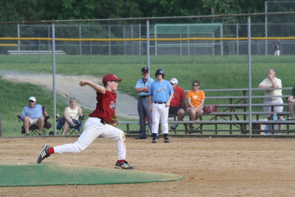 Baseball player pitching with another person on first base and families in the stand.