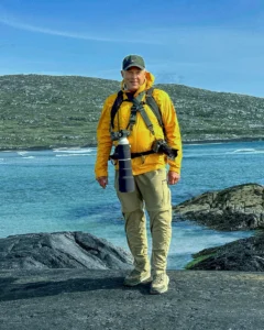 A man with a camera in a yellow jacket and backpack standing in front of a river with a mountain in the background. 