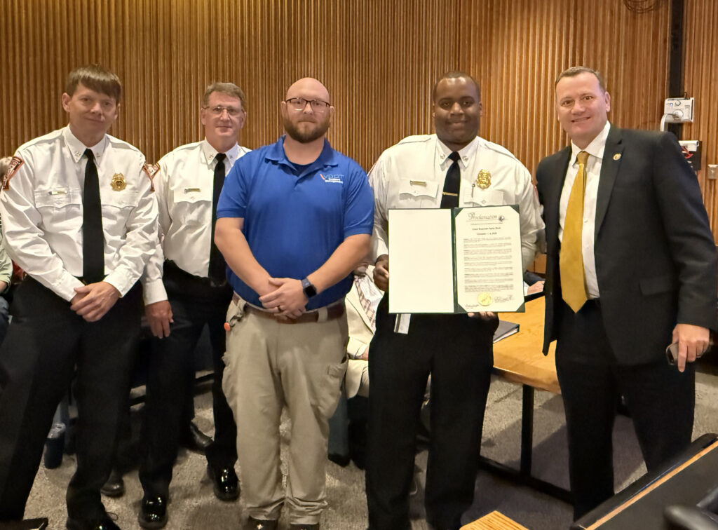 Three firefighters and two others smile for a photo in a meeting room. One holds a proclamation.