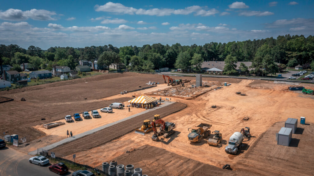 A large construction site with several vehicles, including trucks and construction equipment, is present. There is a yellow and white striped tent in the center. The area is surrounded by trees and nearby houses.