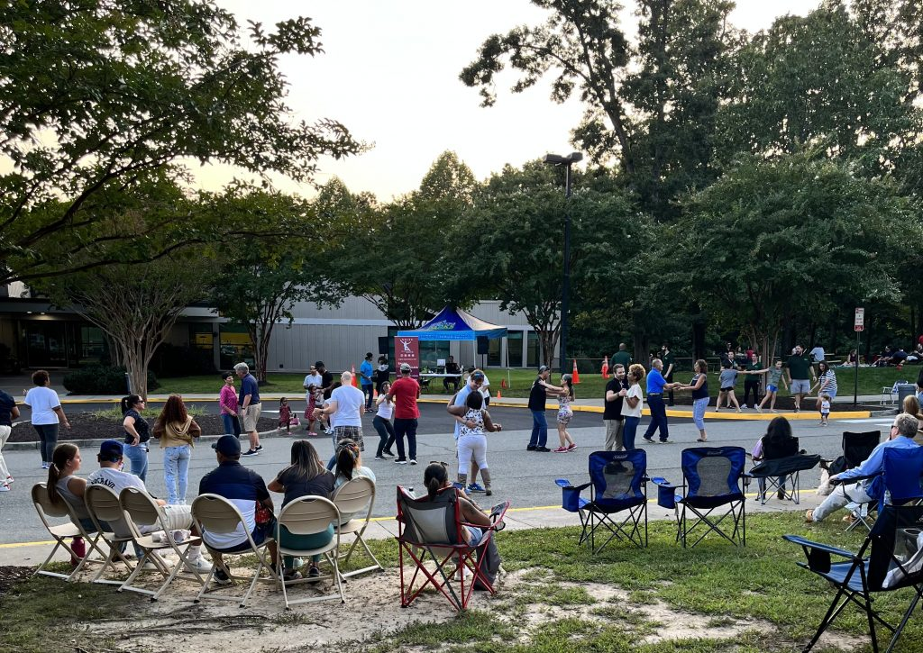 Image: People sitting in lawn chairs outside watching individuals salsa dancing in the parking lot. 