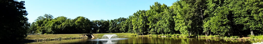 Wide view of a lake and a fountain. The lake is surrounded by a green treeline. Daytime shot.
