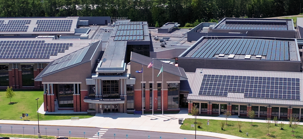 Masthead photo of school of many buildings all with solar panels on the roof. There are 3 flags in front of the building, sidewalks & grass. Photo angle is above the roofs.